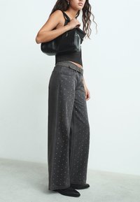 Jeans de mezclilla gris de pierna ancha con detalles de tachuelas, combinados con una camiseta sin mangas negra y un bolso de cuero negro. Textura suave, diseño casual.