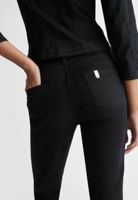 Jeans en denim noir au design ajusté, avec des poches arrière visibles et un accent argenté sur la poche droite. Texture lisse, taille haute.