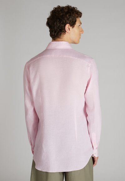 Hombre de espaldas, vistiendo una camisa de botones de manga larga color rosa claro y pantalones verde oliva, frente a un fondo gris liso.