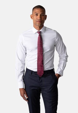 Mann iført hvit skjorte, mønstret rød slips og mørke bukser, står med en hånd i lommen foran ensfarget bakgrunn.