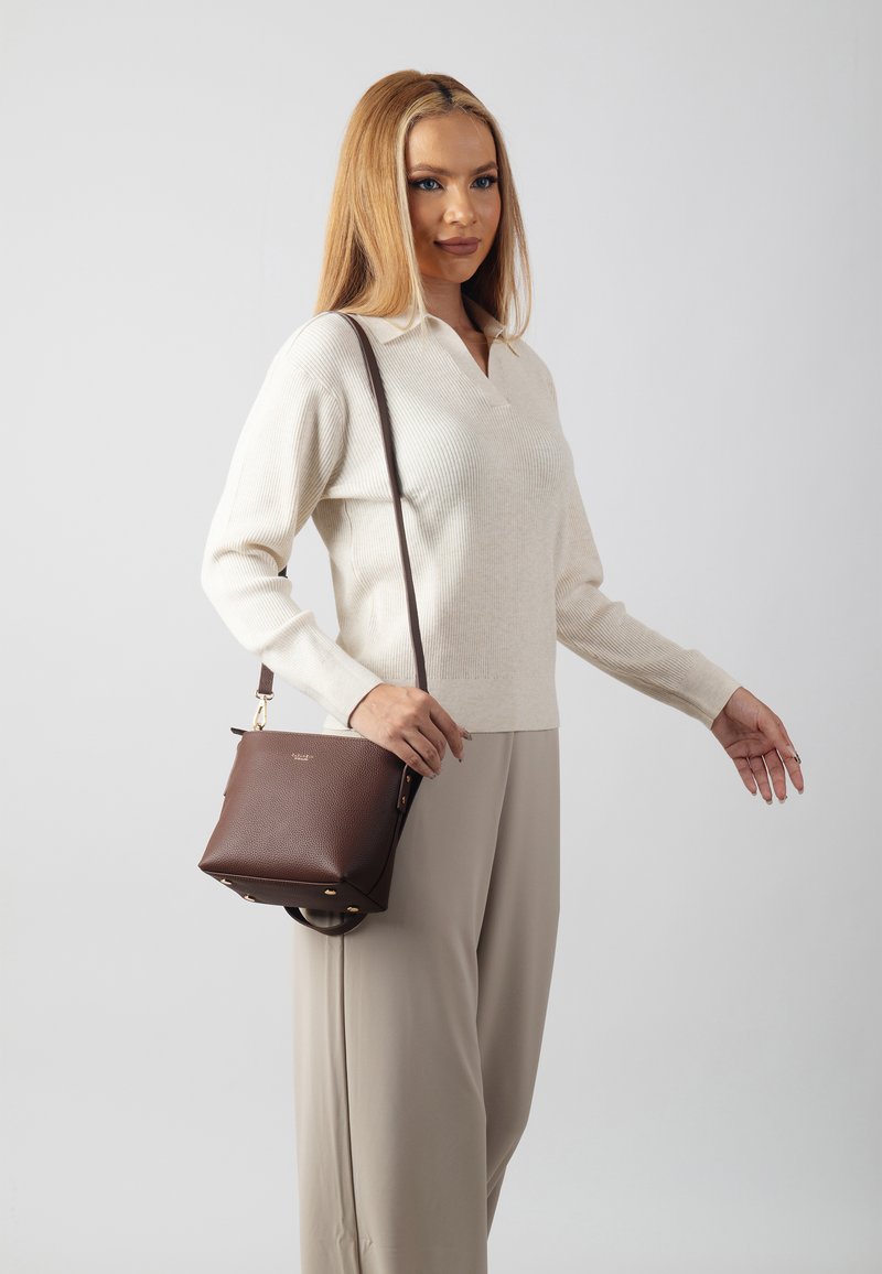 Brown leather crossbody bag with a textured finish, gold hardware, and a strap, paired with a light-colored sweater and beige trousers.