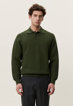 Jeune homme aux cheveux bruns bouclés portant un pull polo texturé à manches longues vert foncé et un pantalon à carreaux foncé sur un fond uni.