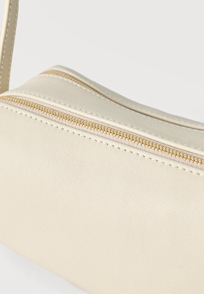 Τσάντα ώμου από κρεμ δέρμα με χρυσή λεπτομέρεια φερμουάρ και εμφανή ράμματα σε απλό λευκό φόντο.