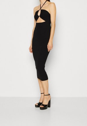 Schwarzes Bodycon-Kleid mit Halterneck, featuring einem V-ausschnitt und einer enganliegenden Silhouette. Kombiniert mit glänzenden schwarzen Plateauschuhen.