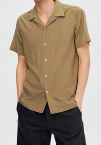 Korta ärmar med krage i olivgrön tyg med subtil lyster. Utrustad med ljusblå knappar och avslappnad passform; matchas med mörka byxor.