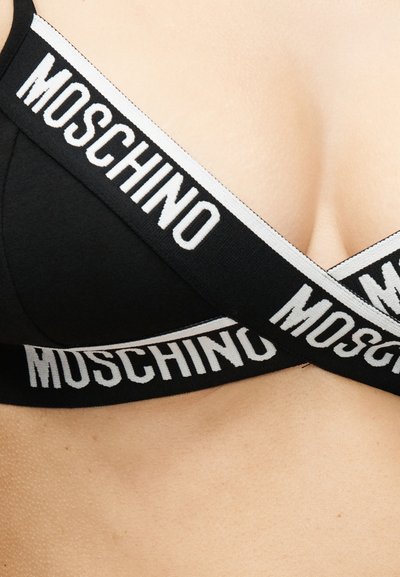 Κοντινό πλάνο ενός μαύρου bralette Moschino με λευκές λωρίδες με λογότυπο που διασταυρώνονται πάνω στο στήθος σε ανοιχτόχρωμο δέρμα.