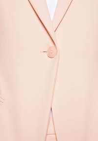 Brzoskwiniowa marynarka z klapą z wycięciem, pojedynczym zapięciem na guzik oraz gładką fakturą. Zawiera subtelny detal przeszycia i przednie kieszenie.