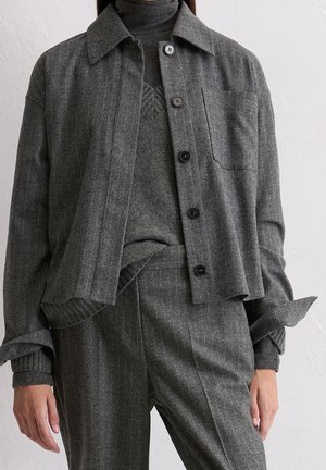 Kvinde iført en grå jakke med knapper over en grå sweater og matchende grå bukser står op ad en hvid væg.