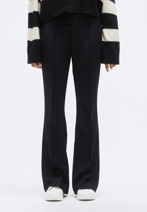 Schwarze, maßgeschneiderte, weit geschnittene Hose mit glatter Textur, vorne mit Bügelfalten und Gürtelschlaufen, kombiniert mit weißen Sneakers.