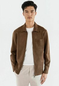 Ung man iklädd en brun zip-up jacka över en vit skjorta och ljusbeiga byxor, stående mot en enfärgad ljus bakgrund.