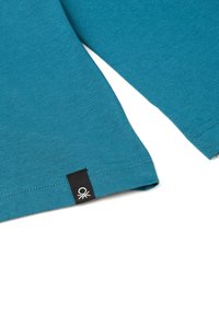 T-shirt in cotone teal con maniche corte e orlo dritto. Presenta un'etichetta nera con un logo bianco in basso a sinistra.