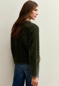 Vrouw met lang bruin haar, gekleed in een donkergroene, zachte trui en een blauwe spijkerbroek, licht zijwaarts gedraaid tegen een effen lichte achtergrond.