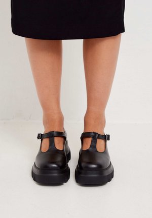 Jambes portant des chaussures Mary Jane noires à semelles épaisses et à brides en forme de T, debout sur un sol clair contre un mur uni.