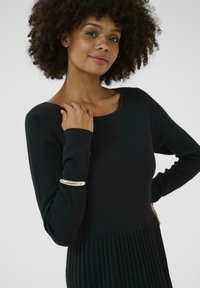 Melns adīts kleita ar gludu augšējo daļu un plisētu apakšējo pusi. Garas piedurknes ar sudrabainu akcentu uz rokas. Mīksta tekstūra.