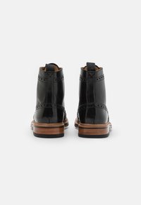 Botas de tobillo de cuero negro con un intrincado detalle de brogue, un tacón de madera en contraste y una lengüeta para facilitar su colocación. Acabado suave y brillante.