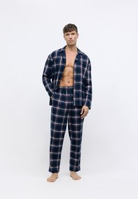 Conjunto de pijama a cuadros azul oscuro con detalles en rojo y blanco. Camisa de manga larga y pantalones con cintura elástica. Material de algodón. Modelo de pie descalzo.