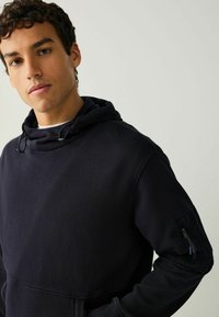 Must kapuutsiga dressipluus tekstuurse viimistlusega, millel on pael, küljetasku ja tõmblukuga varrukadetail.