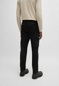 Pantaloni cargo neri con tasche laterali, realizzati in tessuto resistente. Presentano una vestibilità comoda e sono abbinati a una camicia beige a maniche lunghe e a scarpe nere.