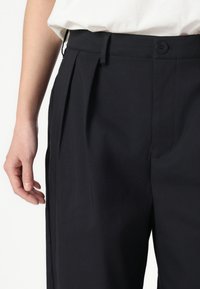 Pantalons plissés noirs avec une texture lisse, présentant une fermeture à bouton et des poches latérales, montrés avec une main reposant sur le tissu.