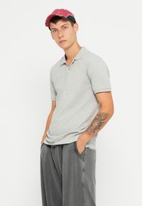 Polo gris à manches courtes, en tissu texturé, avec une patte de boutonnage à trois boutons, associé à un pantalon ample gris foncé et une casquette rouge.