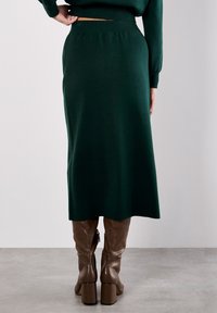 Jupe midi en tricot vert avec une taille ajustée, texture lisse et légère évasement à l'ourlet, associée à des bottes marron jusqu'aux genoux.