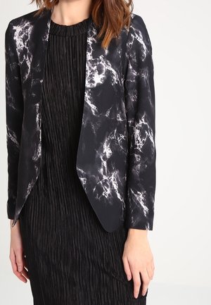 Kvinde iført en sort og hvid blazermønstret med marmorprint over en sort plisseret kjole, stående mod en ensfarvet hvid baggrund.