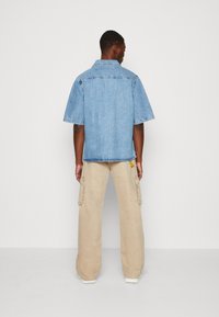 Gai zils īsām piedurknēm džinsu krekls ar taisnu piegriezumu; bēšas krāsas cargo bikses ar sānu kabatām; baltas snikeru kurpes. Neitrāla fona.