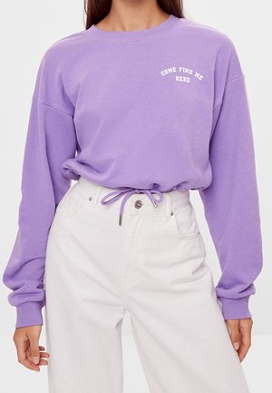 Kvinde iført en lilla langærmet sweatshirt med teksten "COME FIND ME HERE" og hvide bukser med høj talje med et lilla snørebånd som bælte.