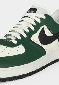 Sneaker Nike basso con pannelli in pelle verde e bianca, lacci bianchi, logo Nike nero e suola in gomma nera.
