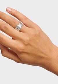 Anillo plateado en forma de ola en una mano. Superficie pulida con secciones alternas abiertas y cerradas. Textura suave. Tono de piel visible debajo.