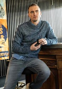 Tjock marinblå stickad tröja med hög krage, lager på lager över en ljusblå skjorta. Slimmade grå jeans och ett mörkt föremål i handen, sittande mot en bar.