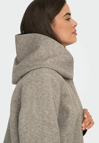 Světlě šedá oversized mikina s velkým límcem, strukturovanou tkaninou a útulným designem. Obsahuje boční kapsy a jednoduchý silueta.
