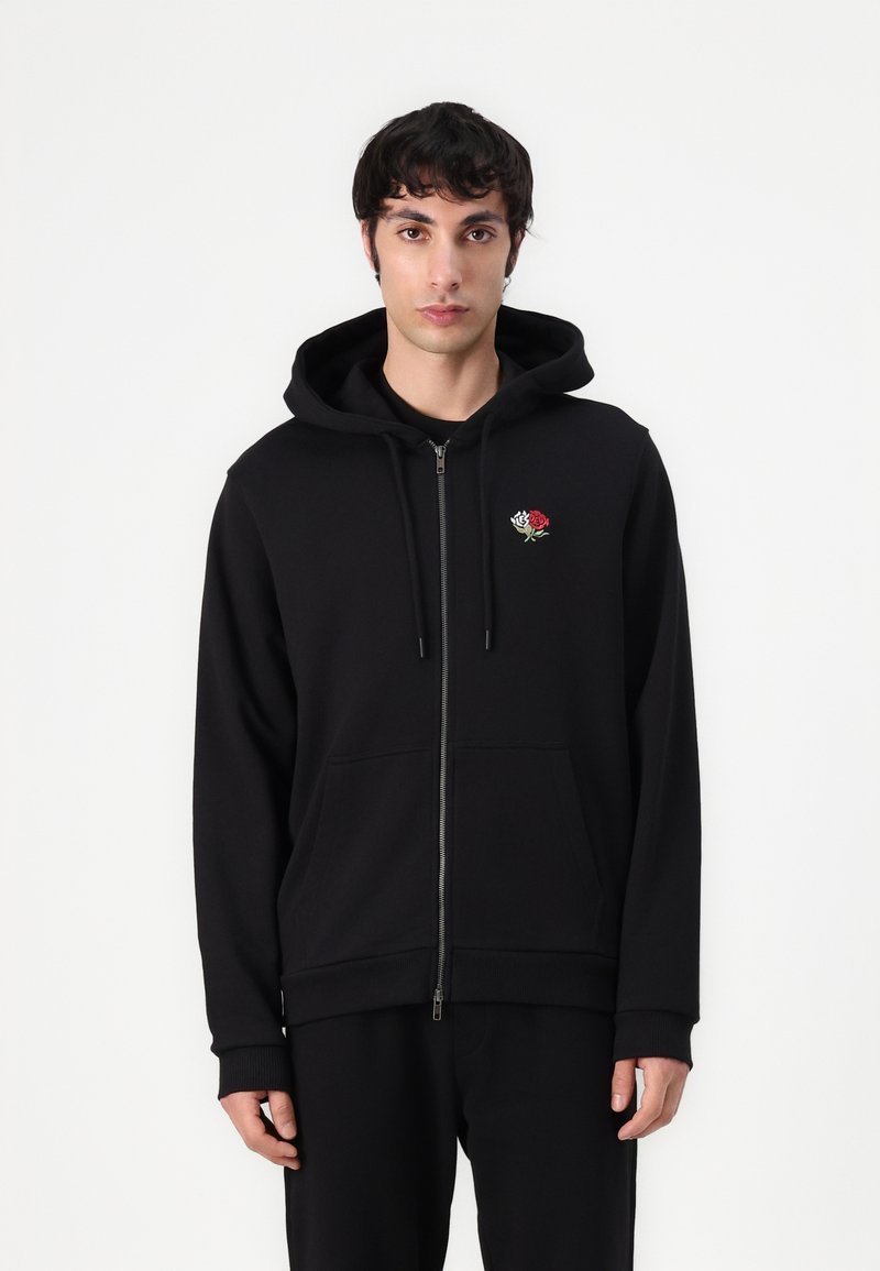 Sudadera con cremallera negra hecha de una tela suave, con capucha, bolsillos frontales y un detalle bordado de una rosa en el lado izquierdo del pecho.