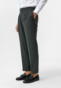Grijze pinstripe broek met zijzakken, geplooid ontwerp en omslagzoom, gecombineerd met zwarte loafers. Textuurdetails van de stof zijn zichtbaar.