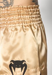 Satin-Shorts in einem hellbeigen Farbton mit einem gerafften, elastischen Bund. Mit einem schwarzen, gestickten Emblem nahe dem Saum. Glatte Textur.