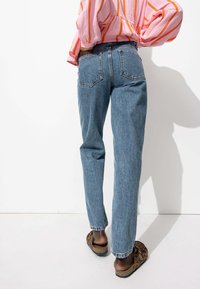 Jean en denim bleu clair avec une coupe droite, comportant deux poches arrière et des détails de couture subtils, portés avec des chaussures à enfiler à motifs brunes.