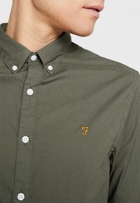 Ciemnozielona koszula z bawełny, z kołnierzykiem zapinanym na guziki, białymi guzikami oraz małym żółtym haftowanym logo na piersi.