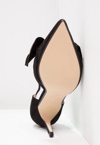 Chaussure à talon haut en daim noir à bout pointu vue de la semelle avec un talon fin reposant contre un mur et un sol blancs.