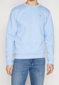 Homme portant un sweat-shirt à manches longues bleu clair avec un petit logo sur la poitrine, associé à un jean bleu délavé moyen, se tenant devant un fond neutre.