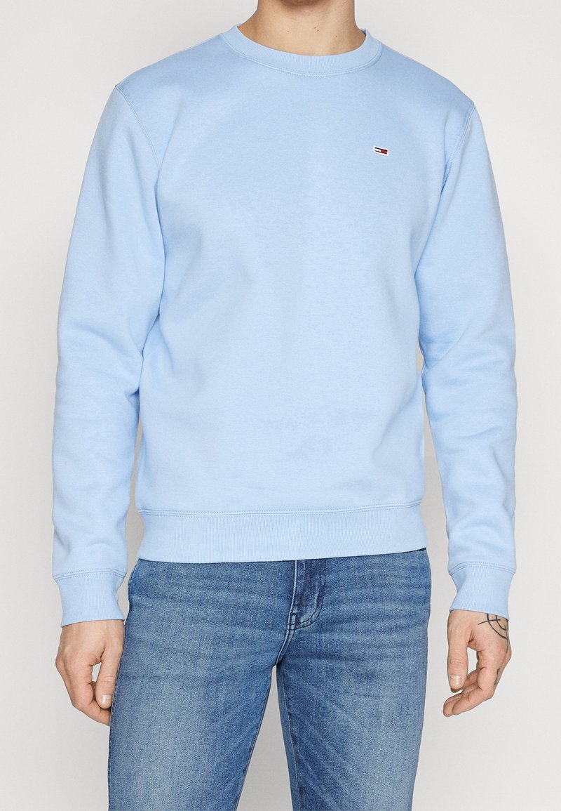 Homme portant un sweat-shirt à manches longues bleu clair avec un petit logo sur la poitrine, associé à un jean bleu délavé moyen, se tenant devant un fond neutre.