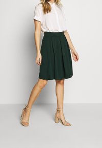 Donna che indossa una blusa bianca a maniche corte con bottoni, una gonna plissettata verde scuro al ginocchio e sandali con tacco a blocco e cinturino alla caviglia color beige.