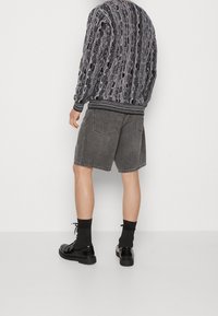 Schwarze und graue gemusterte Jacke mit gerippten Bündchen und Saum, kombiniert mit grauen Jeansshorts und schwarzen Schuhen, von hinten betrachtet.