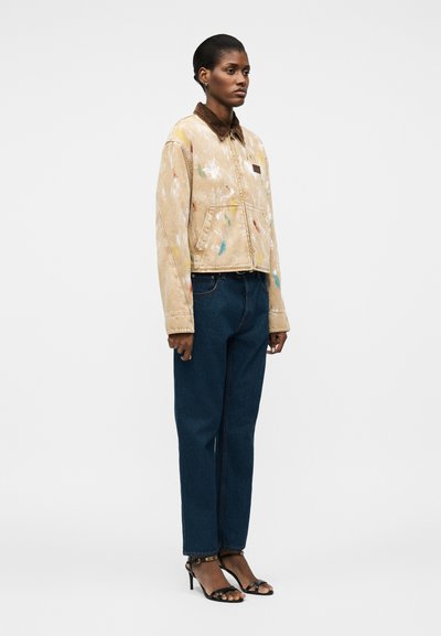 Γυναίκα στέκεται φορώντας ένα μπεζ μπουφάν με πιτσιλιές μπογιάς και καφέ γιακά, σκούρο μπλε τζιν και μαύρα πέδιλα με τακούνι και ανοιχτά δάχτυλα σε λευκό φόντο.