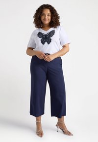 Top blanco de manga corta con un diseño de mariposa de encaje negro, combinado con pantalones de pierna ancha azul marino y tacones plateados con tiras.