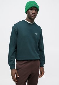 Sötétzöld, kerek nyakú pulóver Calvin Klein logóval, barna nadrággal párosítva. Bordázott mandzsetták és puha anyag. Élénk zöld sapka.