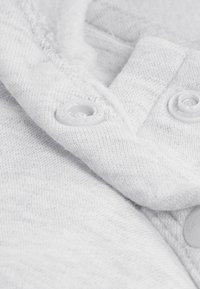 Ανοιχτό γκρι ύφασμα με μαλακή υφή, με λευκά πλαστικά κουμπιά και ορατό ράψιμο κατά μήκος των άκρων. Κοντινή άποψη του υλικού.