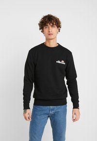 Sudadera negra de manga larga con puños acanalados, que presenta un logo blanco en el pecho y un cuello redondo clásico. Vaqueros de mezclilla visibles por debajo.
