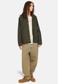 Dunkelgrüne Jacke mit Druckknöpfen, zwei Fronttaschen, getragen über einem hellbeigen Hemd; kombiniert mit lockeren beigen Jeans und braunen Schuhen.