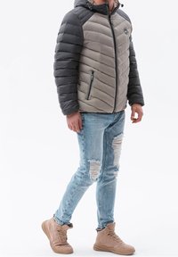 Muž ve šedé a černé padded bundě, roztrhaných světlomodrých džínách a béžových vysokých teniskách, stojící s jednou nohou mírně zdviženou.
