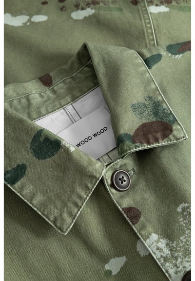 Camisa de algodón verde con cierre de botones, decorada con manchas de verde oscuro y marrón. La etiqueta muestra la marca "WOOD WOOD" dentro del cuello.